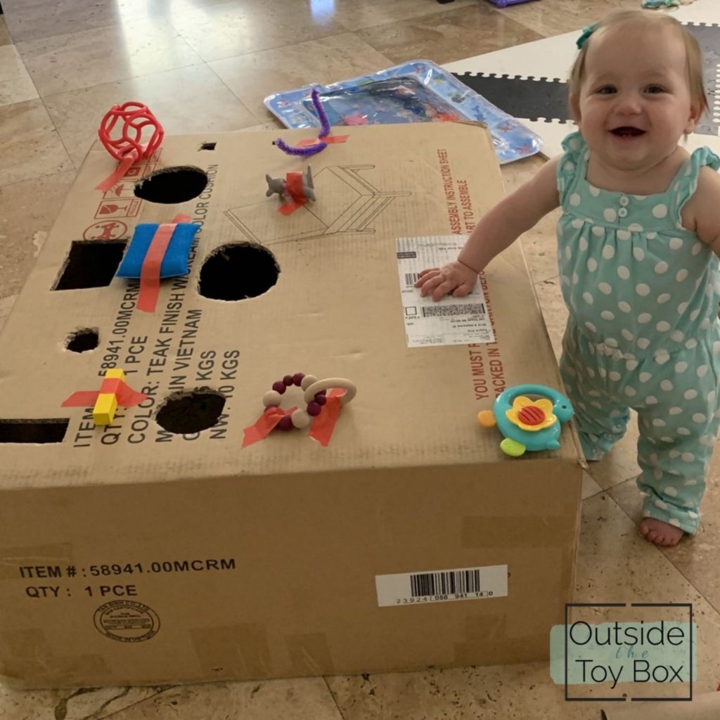 Cardboard box with holes cut out and toys taped on with baby standing holding on