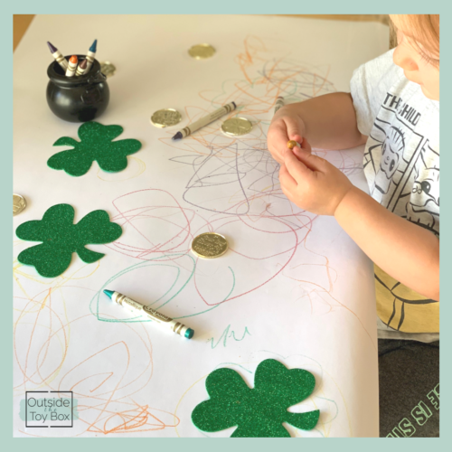 Toddler holding and looking at gold crayon with gold coins and shamrocks on paper