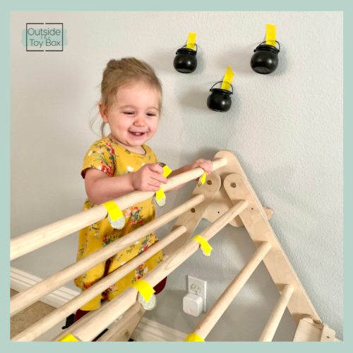 Toddler climbing pikler triangle holding gold coin with pots taped to wall