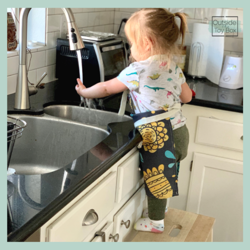 Toddler standing on stool helping clean up paint at the sink